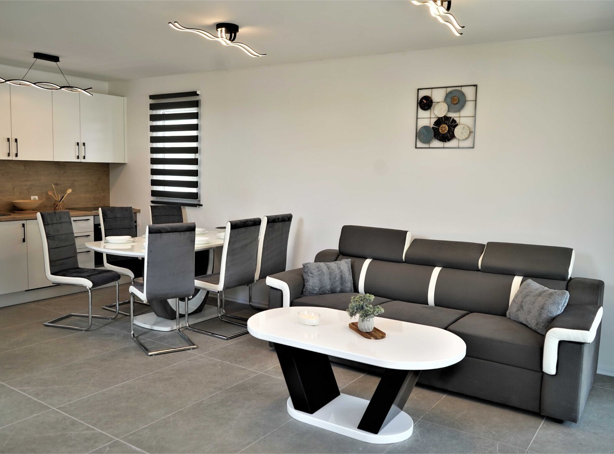 A modern living room with a grey sofa, black and white chairs, a white coffee table, and a kitchen area with cabinets and a countertop.