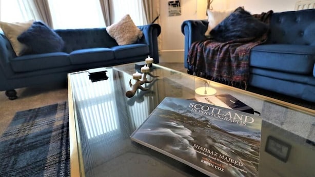 A living room with a glass coffee table, a blue sofa, and a book titled "Scotland in Photographs" by Shahbaz Majeed. - Private 10 Bedrooms Detached Villa in Glasgow | Pool Table | Garden | Piano (Glasgow)