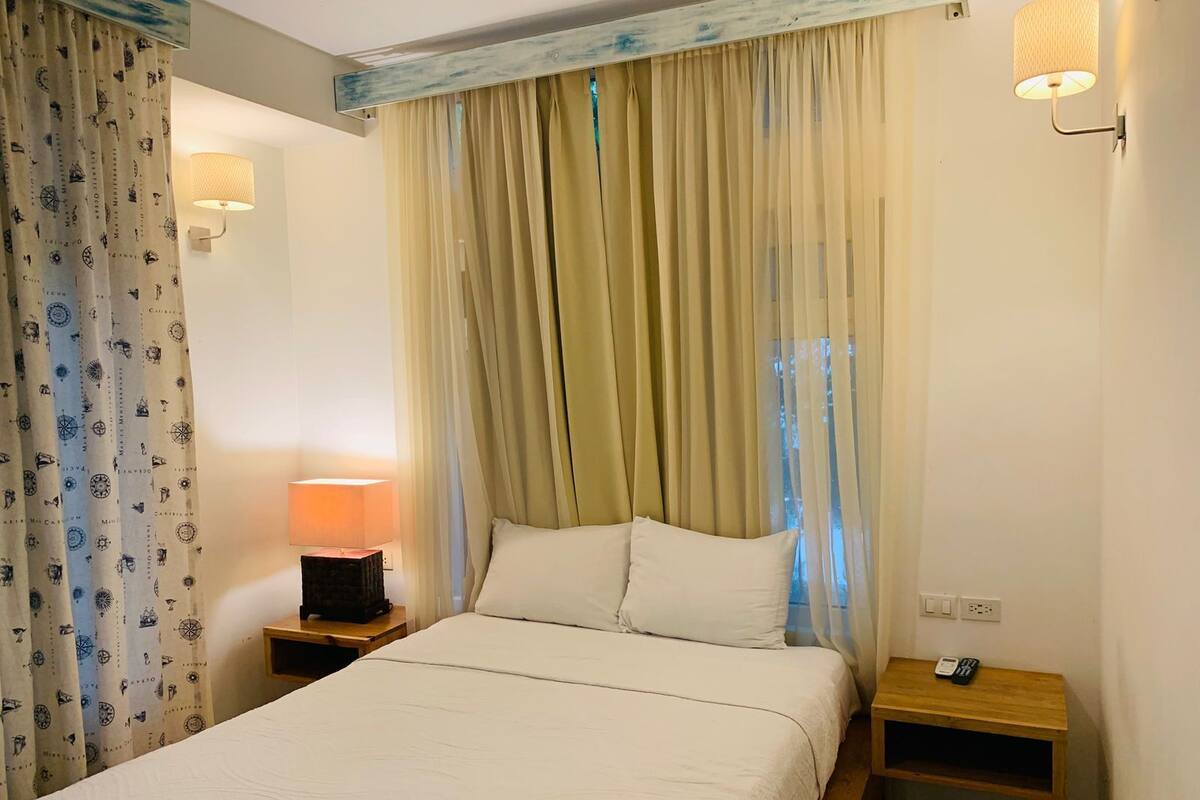 A hotel room with a bed, a nightstand with a lamp, a wooden side table, and a wall-mounted lamp.
