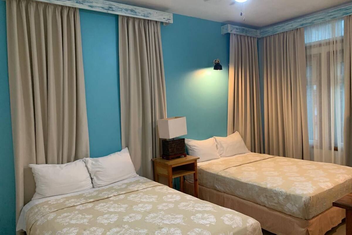 A hotel room with two beds, beige curtains, a nightstand with a lamp, and a ceiling fan.