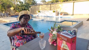 A person pouring a drink into a glass at a poolside table with a bottle of wine, a glass, and a box of wine.