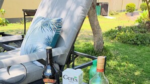 A patio with a lounge chair, a metal table with drinks, a bottle of Bailey's, a box of Ceres orange juice, and a bottle of Captain Morgan.