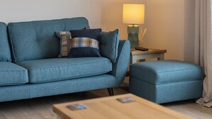 Un salon moderne avec un canapé bleu, un pouf assorti et une table basse en bois. Une lampe est posée sur une petite table d’appoint à côté du canapé.