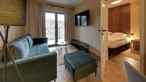 A modern living room with a sofa, ottoman, and a small table. A TV is mounted on the wall, and a bed is visible through an open door. - RentPlanet - Apartament Belle View (Szklarska Poreba)