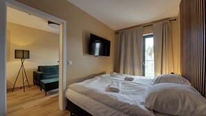 A hotel room with a bed, a TV mounted on the wall, a floor lamp, and a view of the outdoors through the window. - RentPlanet - Apartament Belle View (Szklarska Poreba)