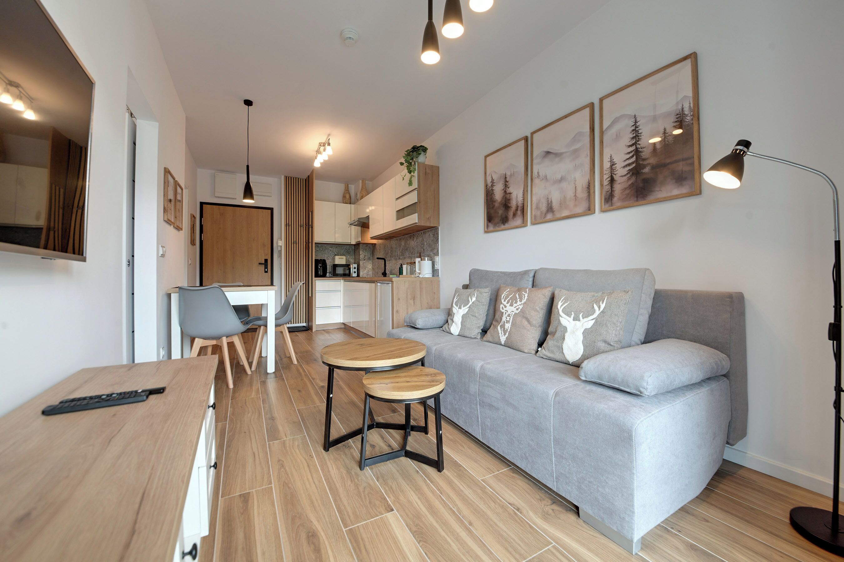 A modern living room with a grey sofa, two round wooden tables, a dining area with a white table and chairs, and a kitchen in the background.