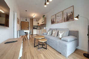 A modern living room with a grey sofa, two round wooden tables, a dining area with a white table and chairs, and a kitchen in the background. - RentPlanet - Apartament Belle View (Szklarska Poreba)