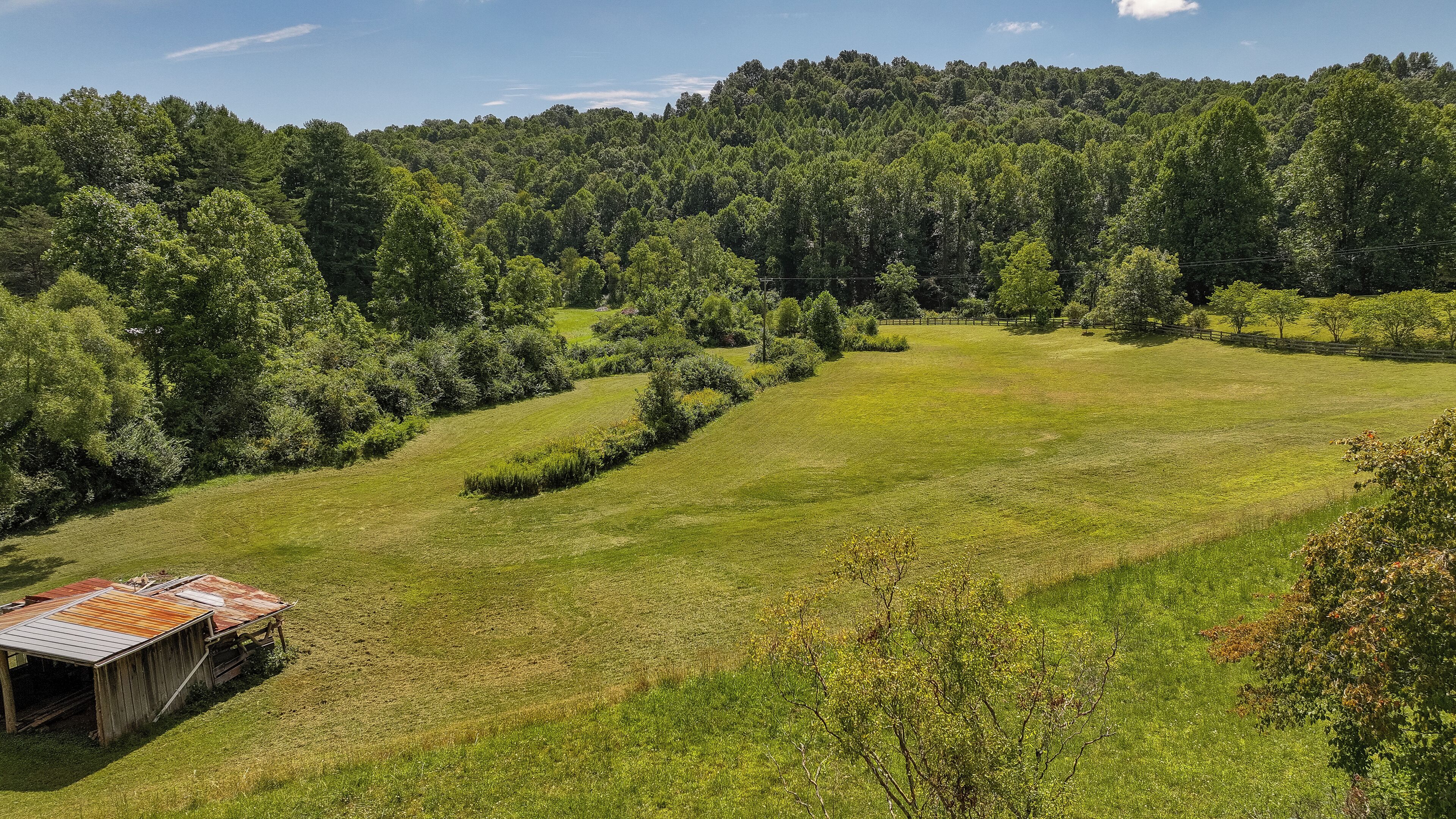 A rural landscape with a grassy field, a wooden structure, and a forested hill in the background.