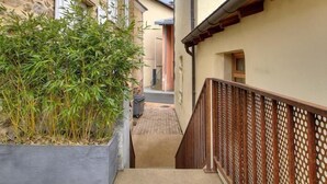 Un estrecho callejón entre edificios, con un macetero de concreto con bambú, una cerca de madera y un edificio de piedra con balcón.