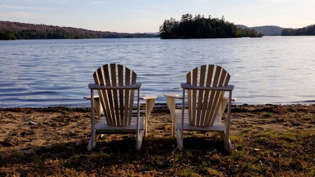 Deux fauteuils Adirondack sont placés face à un lac, avec une île boisée qui se profile à l’horizon.
