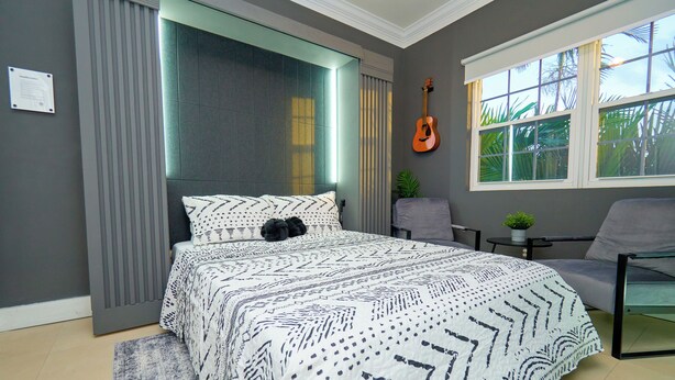 Quarto moderno com uma cama grande, um violão na parede e vista para palmeiras pela janela.