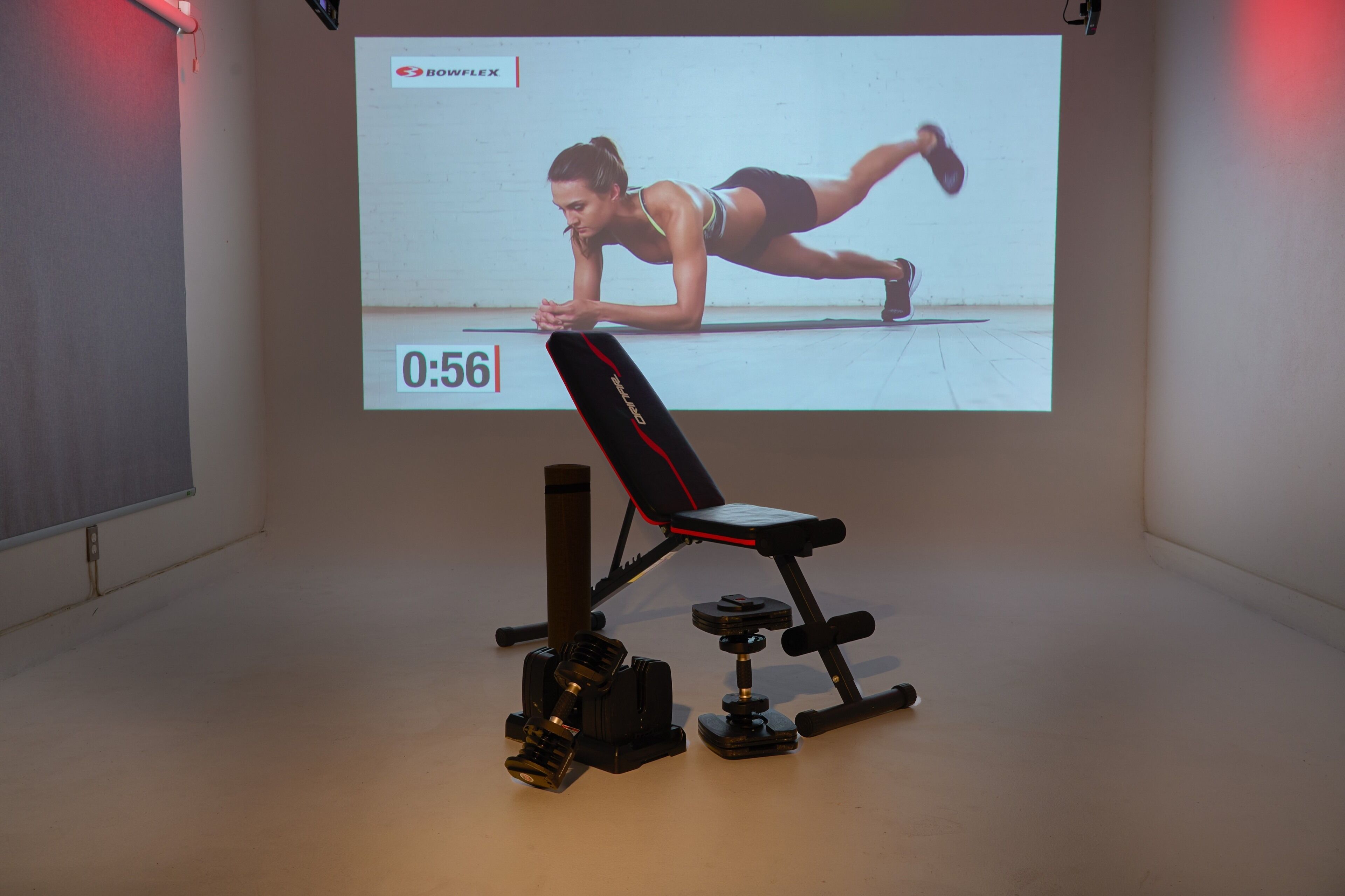 Une salle de sport équipée d’un banc, d  ’haltères et d’un écran de projection diffusant une vidéo d’entraînement physique.