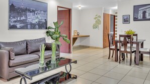 A modern living room with a sofa, coffee table, dining table, and chairs. There are two large paintings on the walls, a potted plant, and a wooden cabinet.