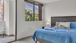 A bedroom with a bed, a bedside table with a lamp, a tray with fruits and drinks, and a view of greenery through the window.