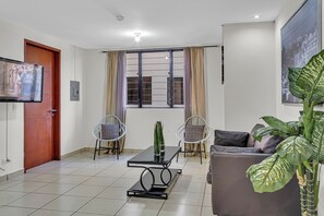Relax in this stylish living room with a flat-screen TV, comfortable sofa, and two chairs, offering a view of the outside through large windows.