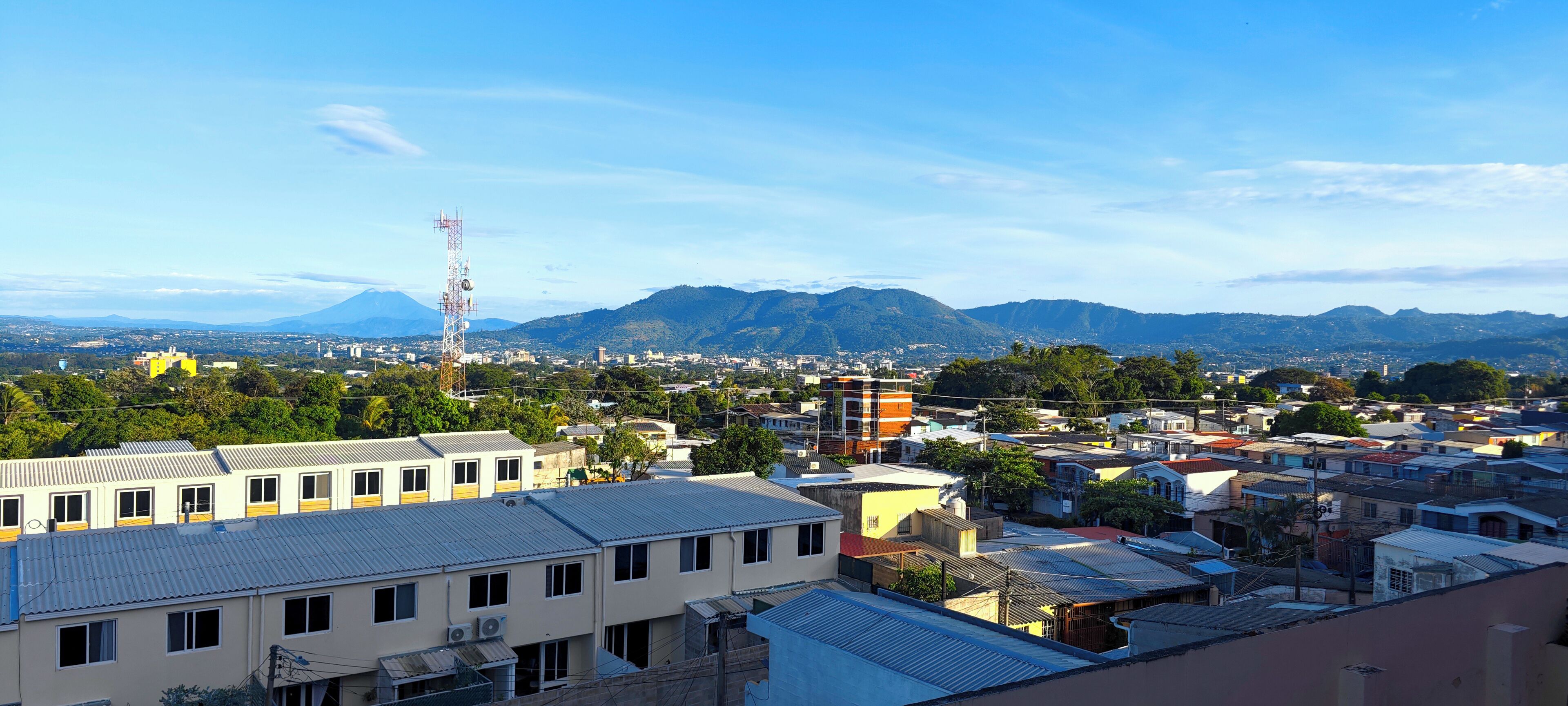 A cityscape with buildings, a prominent communication tower, and mountains in the background.
