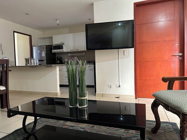 Una sala de estar moderna con una mesa de centro de vidrio, un televisor montado en la pared, una puerta de madera y una cocina al fondo.