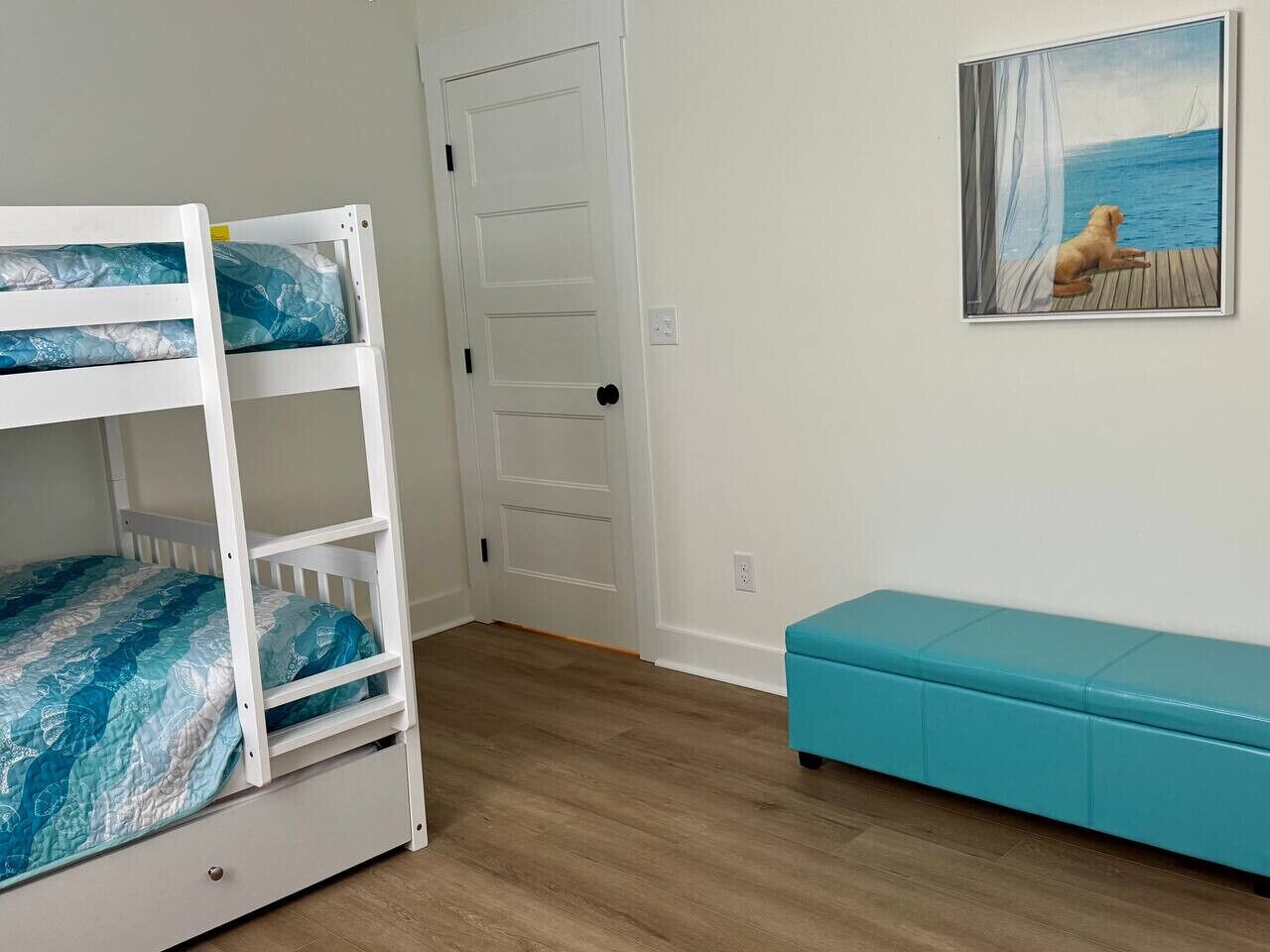 A room with a bunk bed, a door, a bench, and a painting of a dog by the sea.
