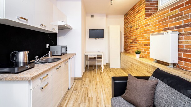Cocina compacta con gabinetes blancos, encimera negra y cafetera. Hay una mesita con una silla, un televisor montado en la pared y una pared con diseño de ladrillo.