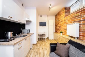 A compact kitchen with white cabinets, a black countertop, and a coffee maker. There is a small table with a chair, a TV mounted on the wall, and a brick-patterned wall.