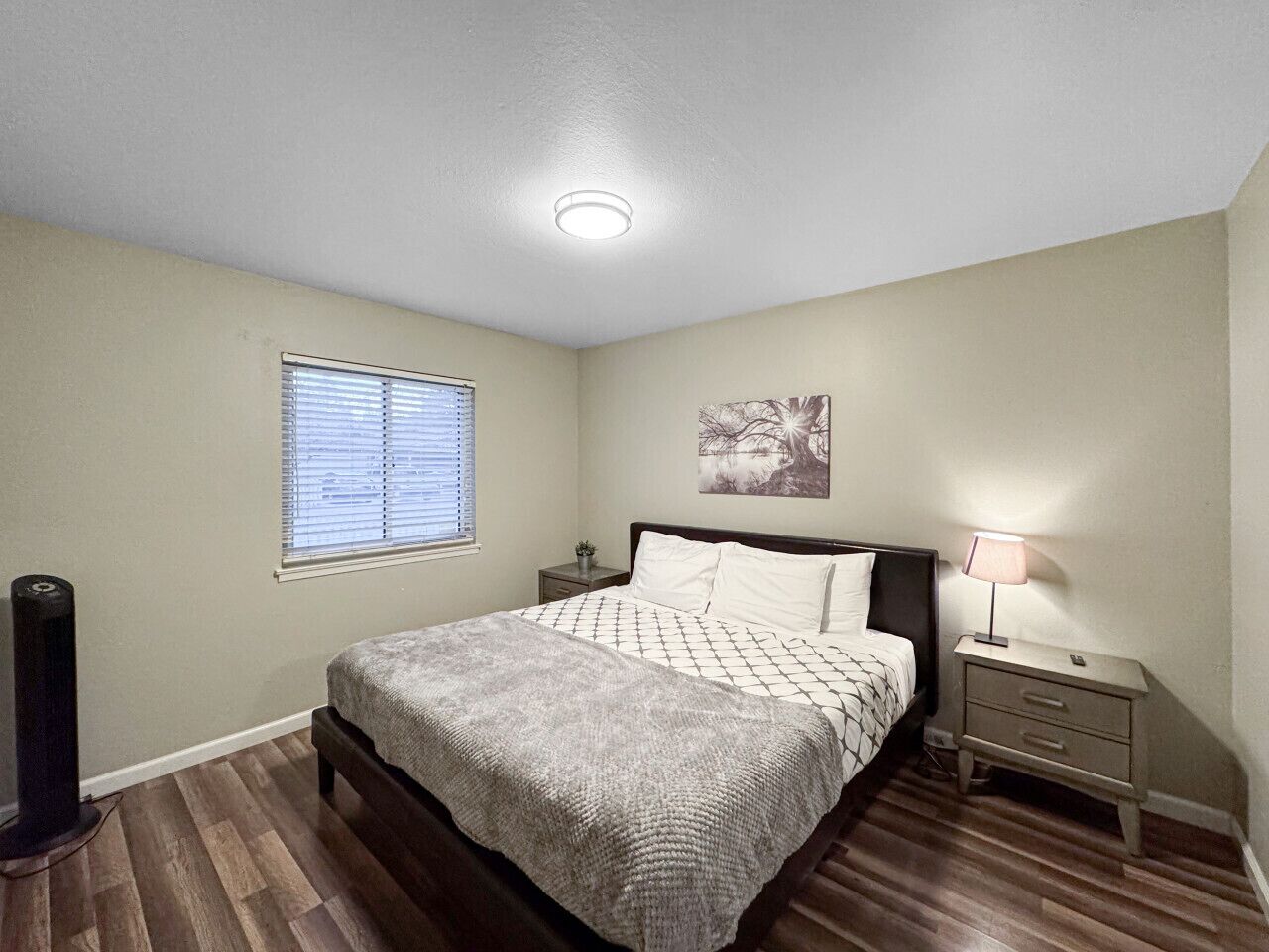 Une chambre à coucher comprenant un lit, des tables de chevet, une lampe, une fenêtre avec des stores et un luminaire au plafond.
