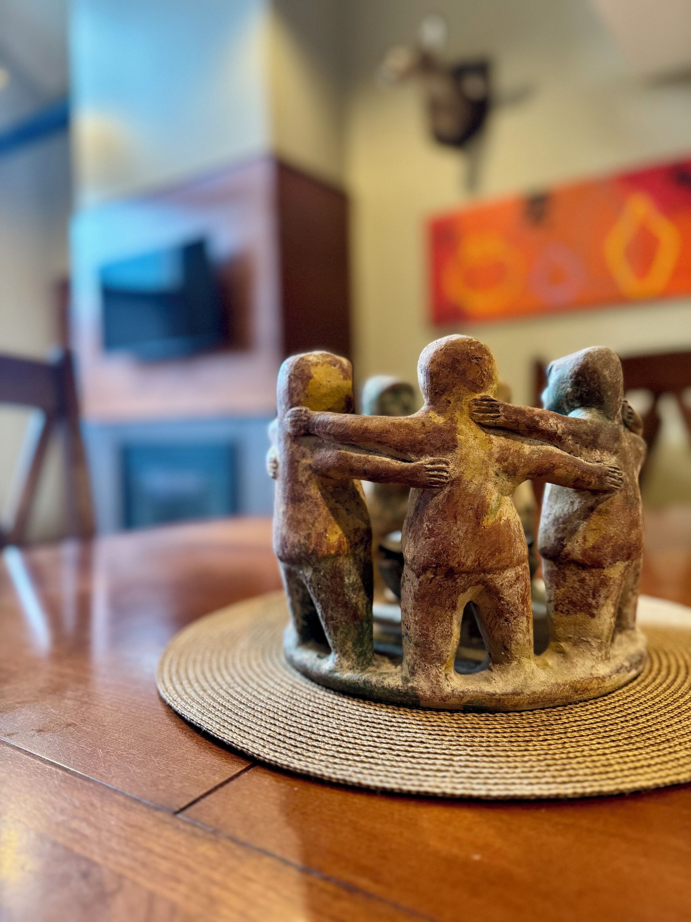 A wooden table with a woven placemat, a small sculpture of four figures holding hands, and a blurred background with a television and a painting.