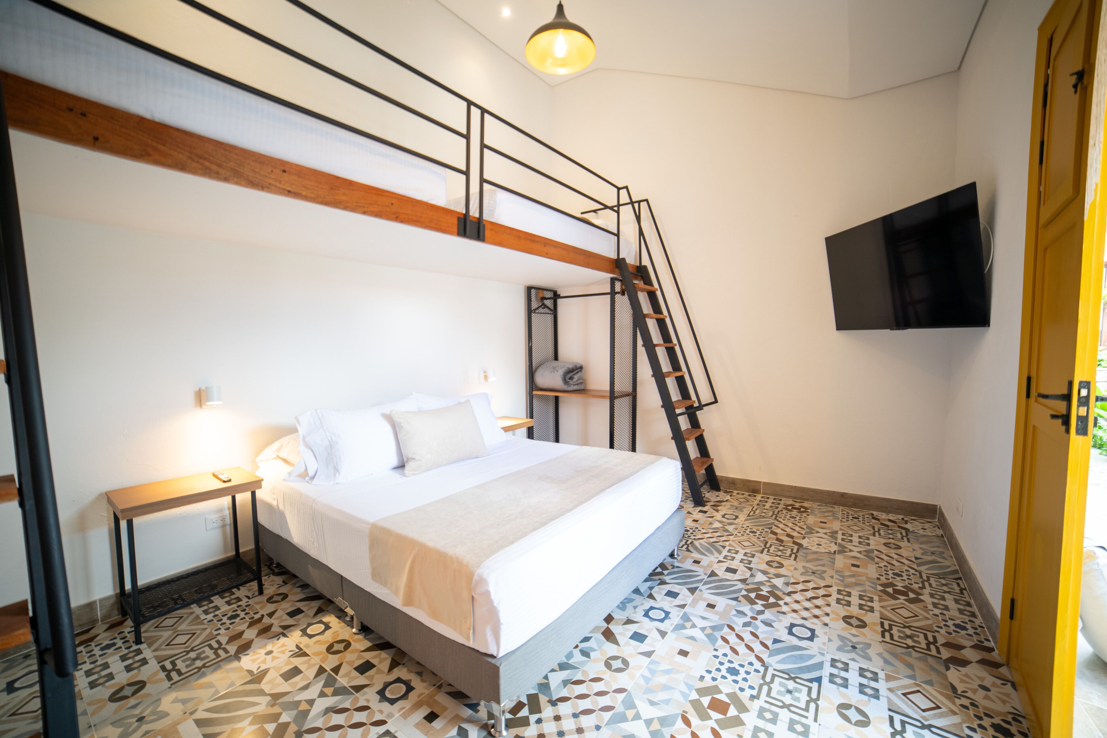 A bedroom with a bunk bed, a ladder, a bedside table with a lamp, and a patterned floor.