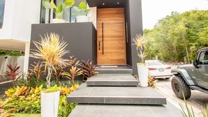 Una casa moderna de dos pisos con puerta principal de madera, un pequeño jardín con diversas plantas y un vehículo estacionado en la entrada.