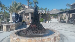 A stone fountain with a central column and surrounding seating area, palm trees, and buildings in the background. - Hacienda La Ballena  (Cabo San Lucas)