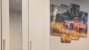 Una habitación con un armario blanco, una cama con cubierta oscura y una pared con una obra de arte de tres paneles que muestra un camión rojo en un campo.