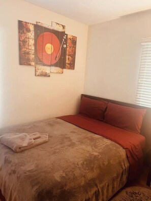 Un lit simple avec une tête de lit rouge et une couverture marron, un oreiller rouge et une œuvre d’art abstraite accrochée au mur.