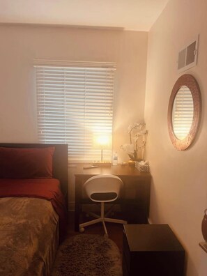 A bedroom with a bed, a desk, a chair, a small table, and a mirror.