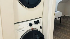 Máquina de lavar e secar empilhadas, com visor digital, em um quarto com teto inclinado e uma cama ao fundo.