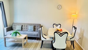 Uma sala de estar moderna com um sofá cinza, uma mesa de centro de madeira e uma mesa de jantar com cadeiras brancas. Um relógio e uma luminária de parede são visíveis.