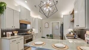 Una cucina moderna con mobili bianchi, un'isola centrale con sottopiatti, piano cottura integrato e una lampada a sospensione geometrica.