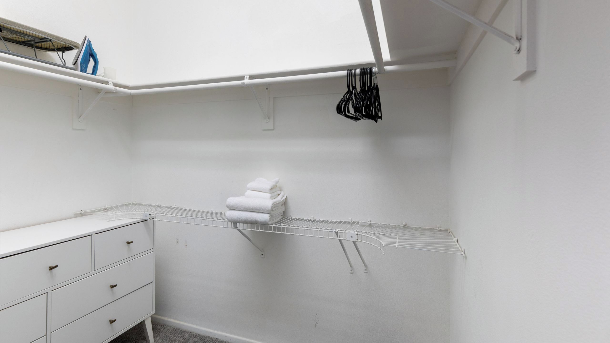 A white closet with a hanging rack, a folded towel on the floor, and a white dresser with drawers.