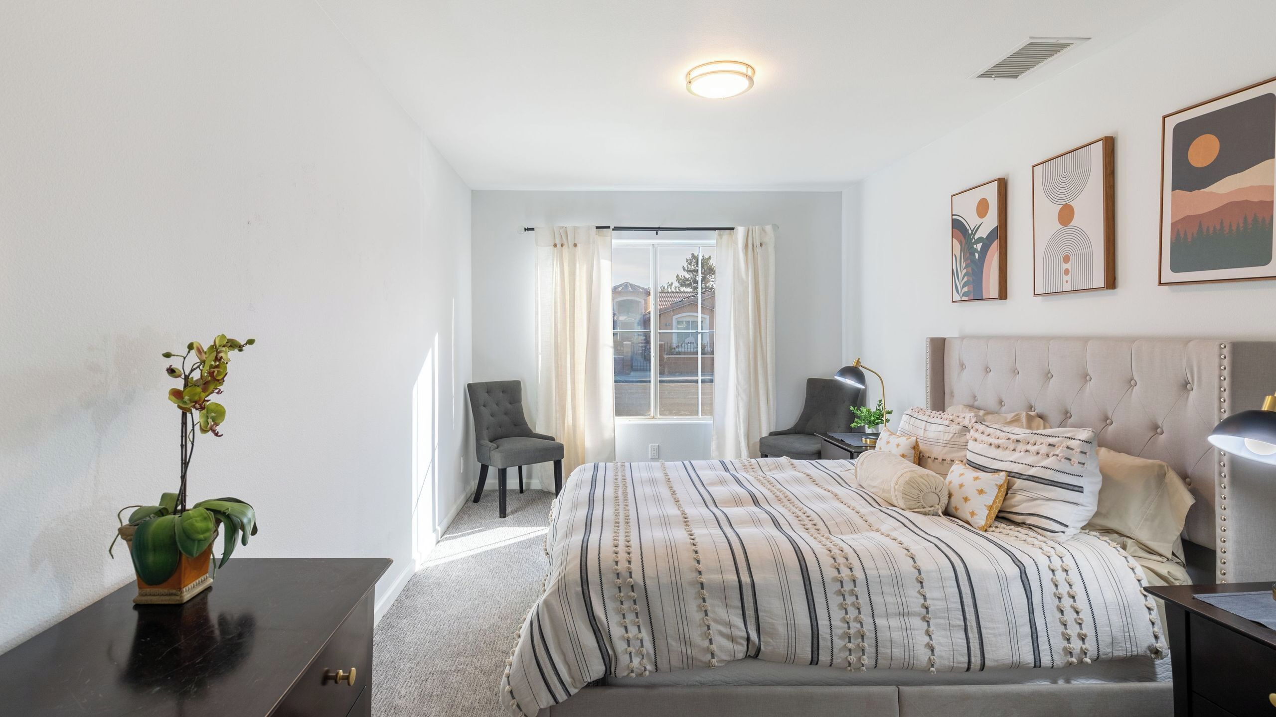 A bedroom with a bed, two chairs, a desk, a lamp, and a potted plant.
