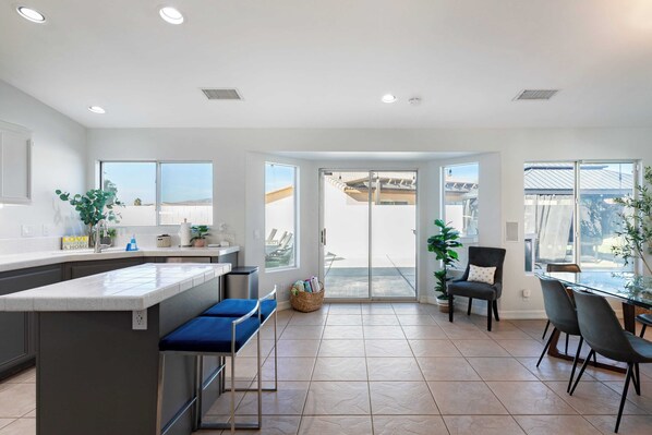 A modern kitchen with a large island, a dining area with chairs, and a view through glass doors to an outdoor patio. - Pacifica Sur by Avantstay Pool, Hot Tub, Gazebo (Indio)