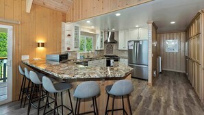 A modern kitchen with wooden paneling, a granite island with bar stools, stainless steel appliances, and a view of greenery outside. - Casa Del Lago Spacious 4 Story Gated Home (Lake Arrowhead)