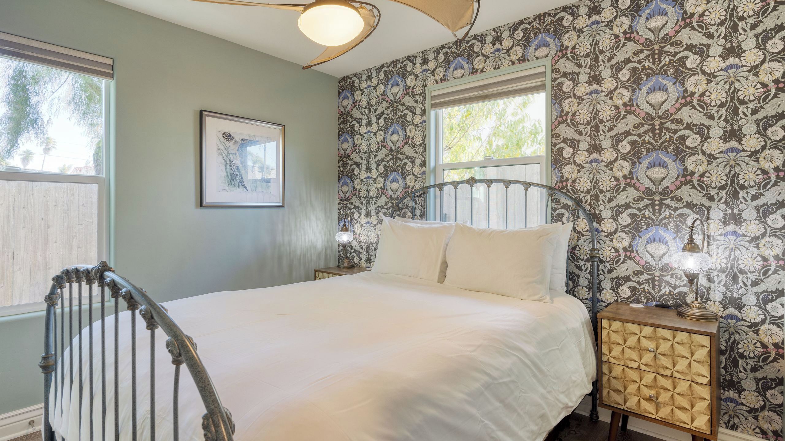 A bedroom with a bed, two bedside lamps, a framed picture on the wall, and a window with a view of trees.
