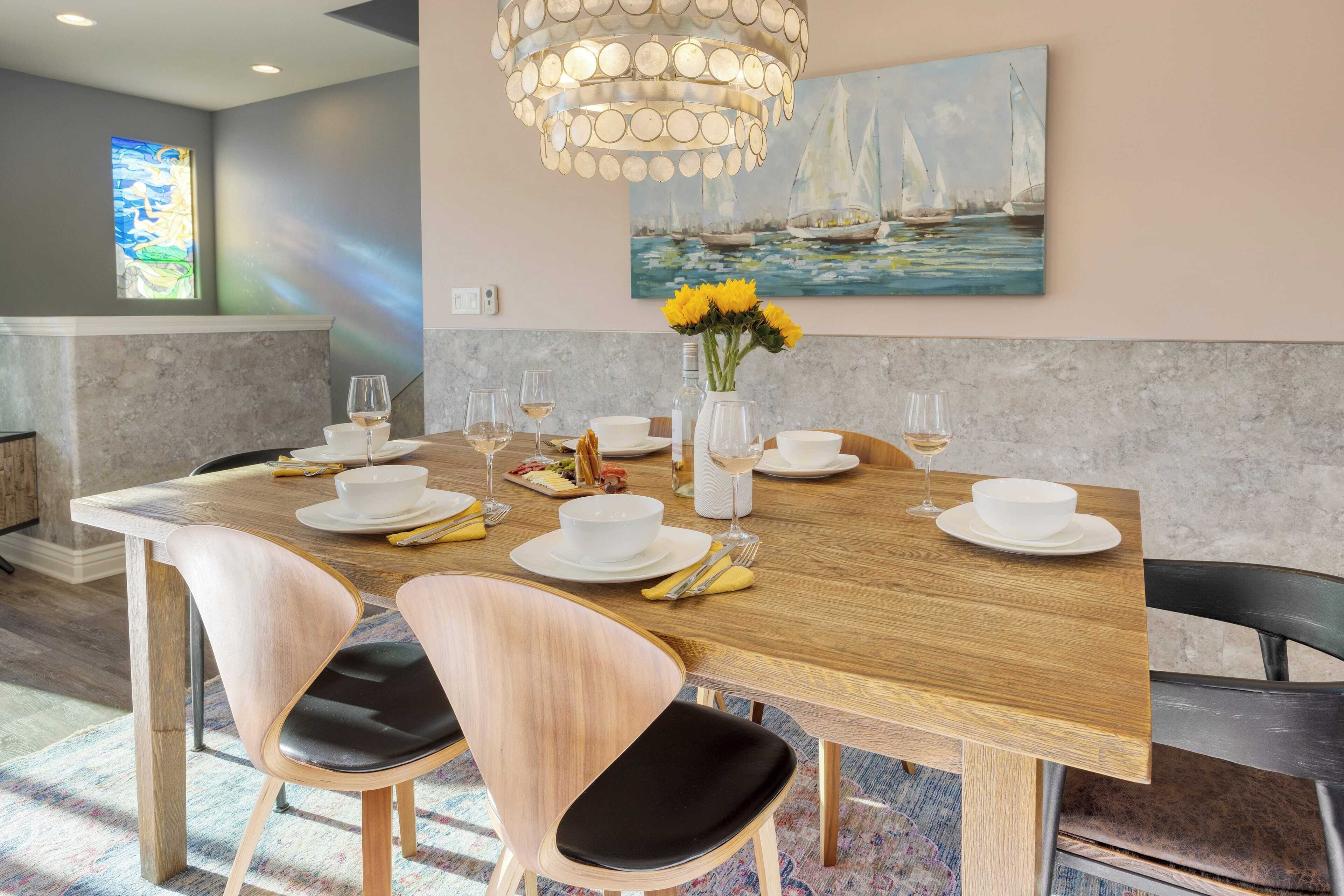 Un coin repas avec une table en bois dressée pour six personnes, entourée de chaises modernes, d’un lustre et d’un tableau représentant des voiliers accroché au mur.