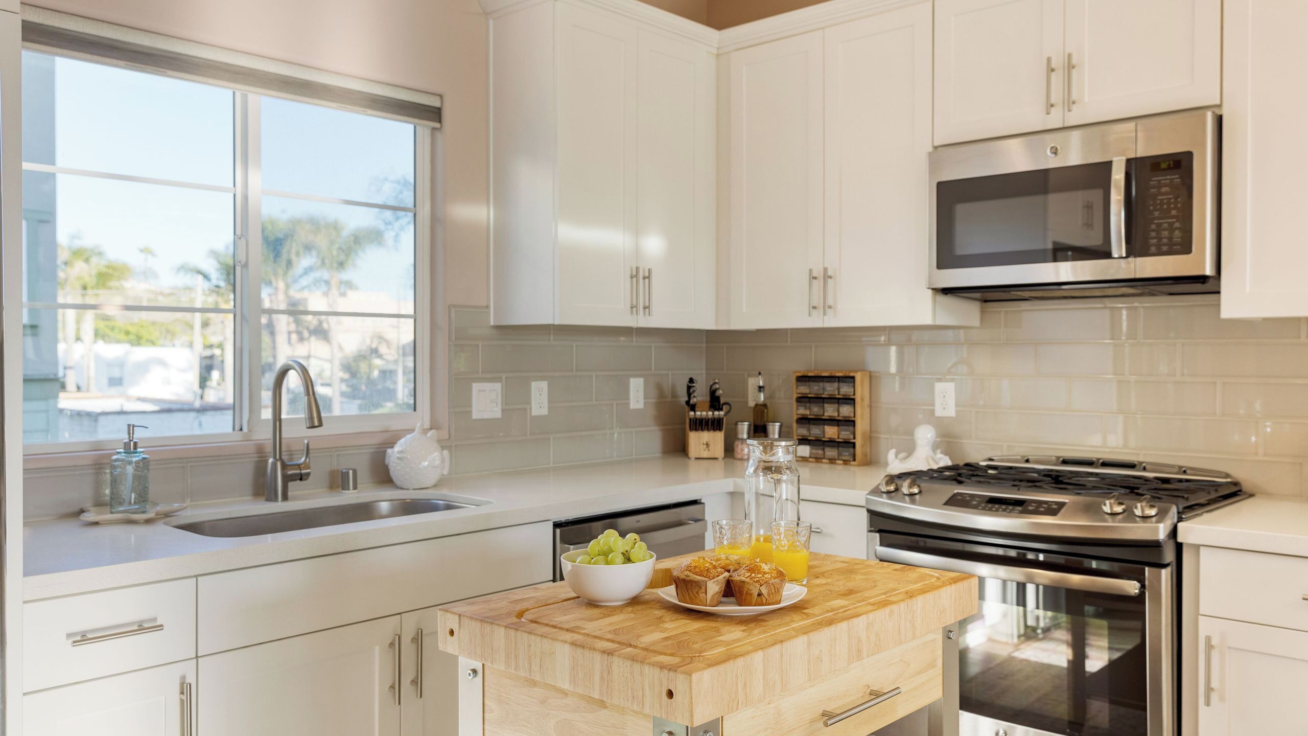 A modern kitchen with white cabinets, a stainless steel stove, a microwave, and a window with a view of palm trees.