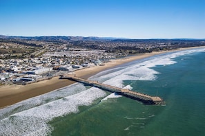 Vista aérea de un pueblo costero con un muelle largo que se adentra en el océano, una playa de arena y una zona residencial densa al fondo.