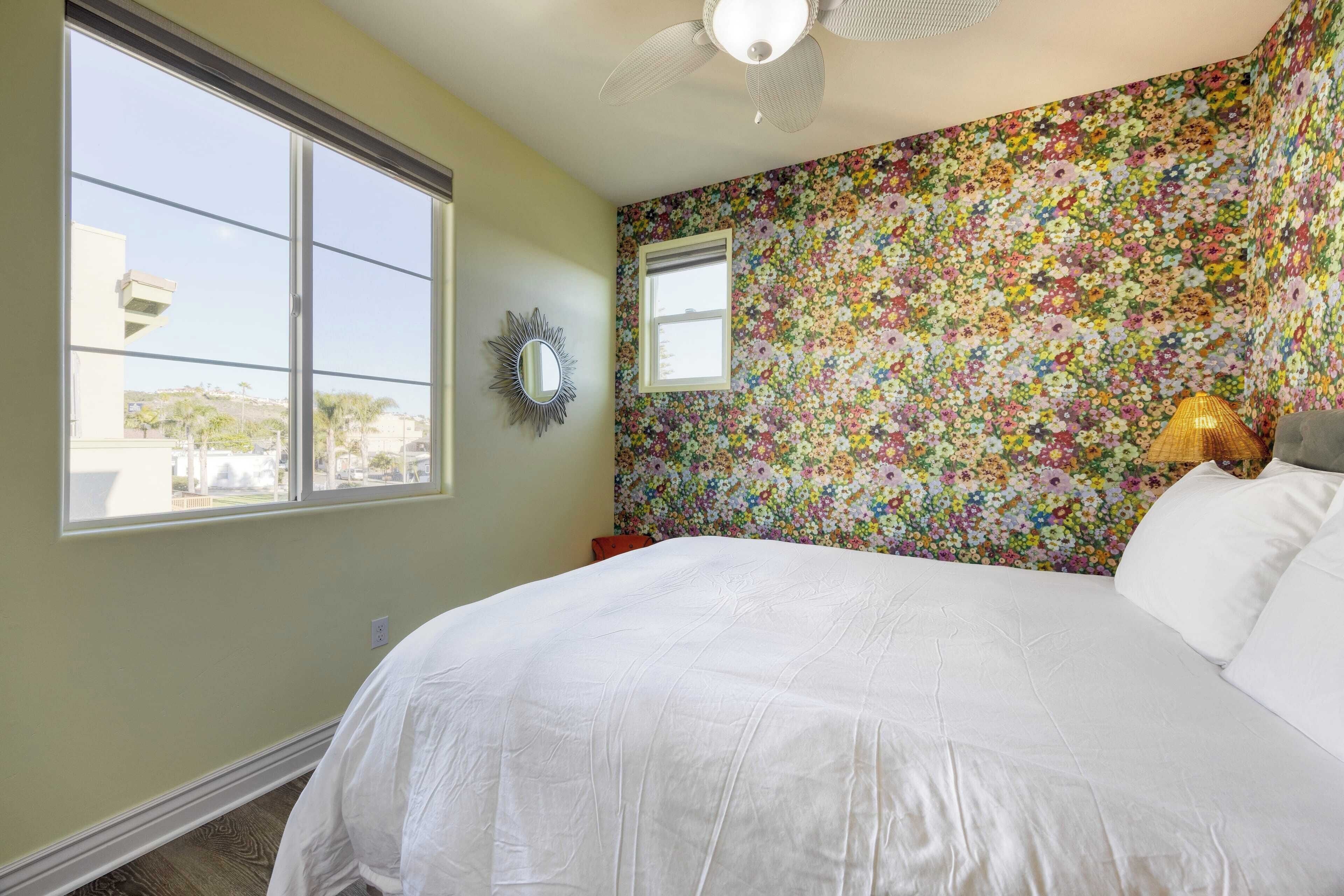 Um quarto com uma cama grande, um ventilador de teto, uma janela e papel de parede floral.