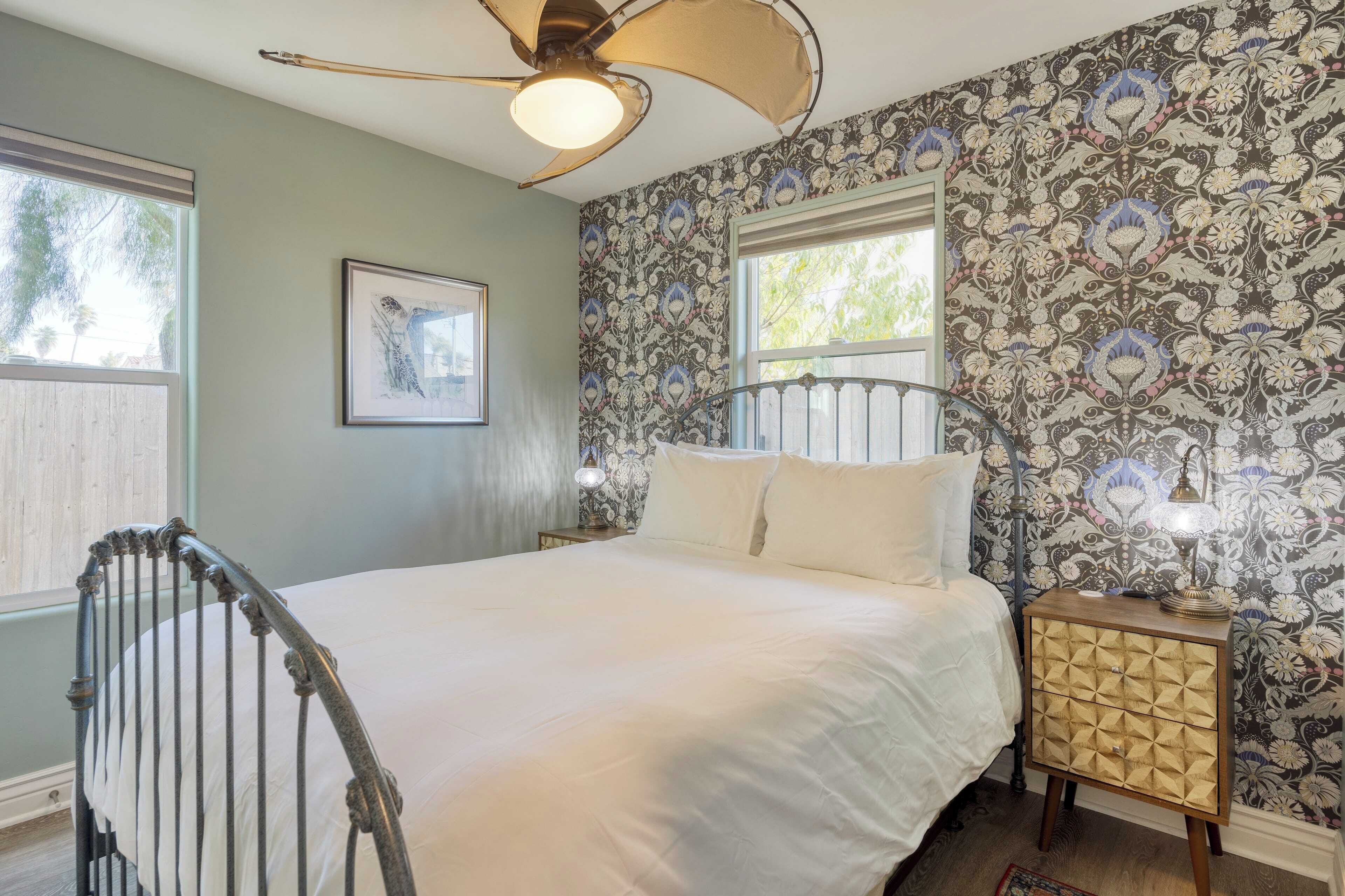 A bedroom with a bed, a nightstand, a chair, a ceiling fan, and patterned wallpaper.