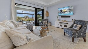 A modern living room with a large TV, comfortable seating, and a view of the outdoors. - Brand New & Luxurious, Sunsets, Heated Pool & Spa - Villa Palm Breeze - Roelens (Cape Coral)