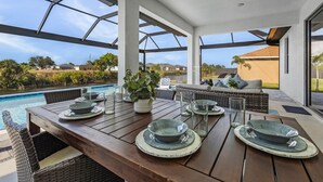 An outdoor dining area with a pool view, wooden tables, and wicker furniture. - Brand New & Luxurious, Sunsets, Heated Pool & Spa - Villa Palm Breeze - Roelens (Cape Coral)