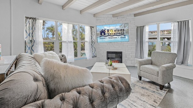 A spacious living room with a fireplace, large windows, and a view of palm trees. - Gulf Access, Dock, Heated Pool, Across from the Beach - Sea-cluded Bungalow -Roelens (Fort Myers Beach)