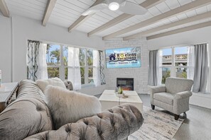 A spacious living room with a fireplace, large windows, and a view of palm trees.