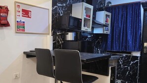 A small kitchenette with a microwave, kettle, and a shelf with kitchen items. There are two black bar stools and a whiteboard with emergency instructions.